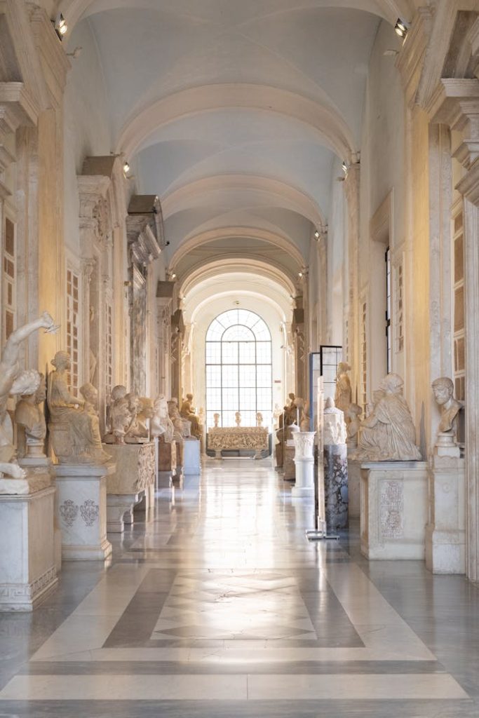 Sunlit hallway featuring classical sculptures in an ornate museum interior.